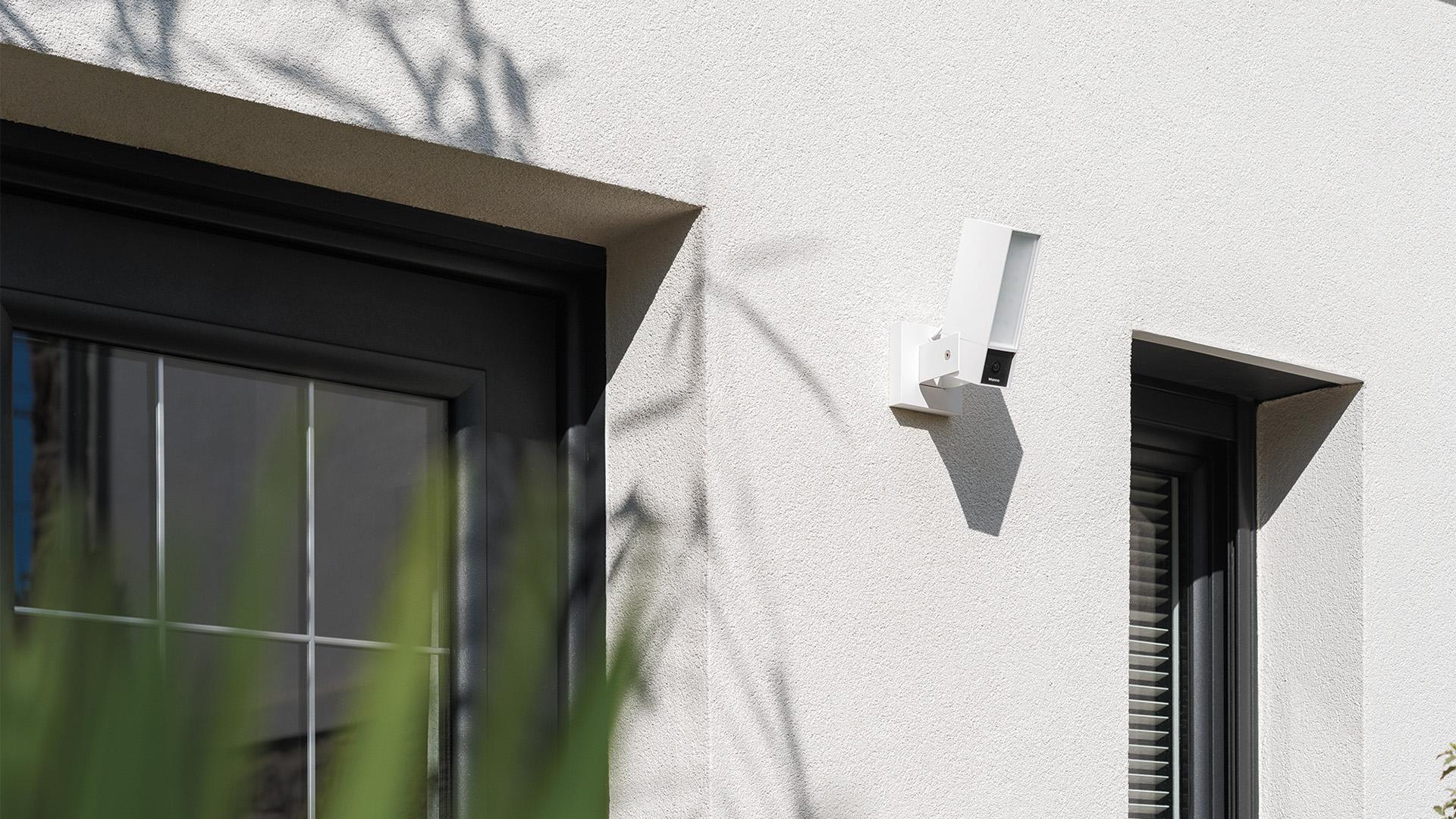 Una telecamera BTicino Outdoor Original bianca è installata all'esterno, tra una porta e una finestra di un edificio moderno.