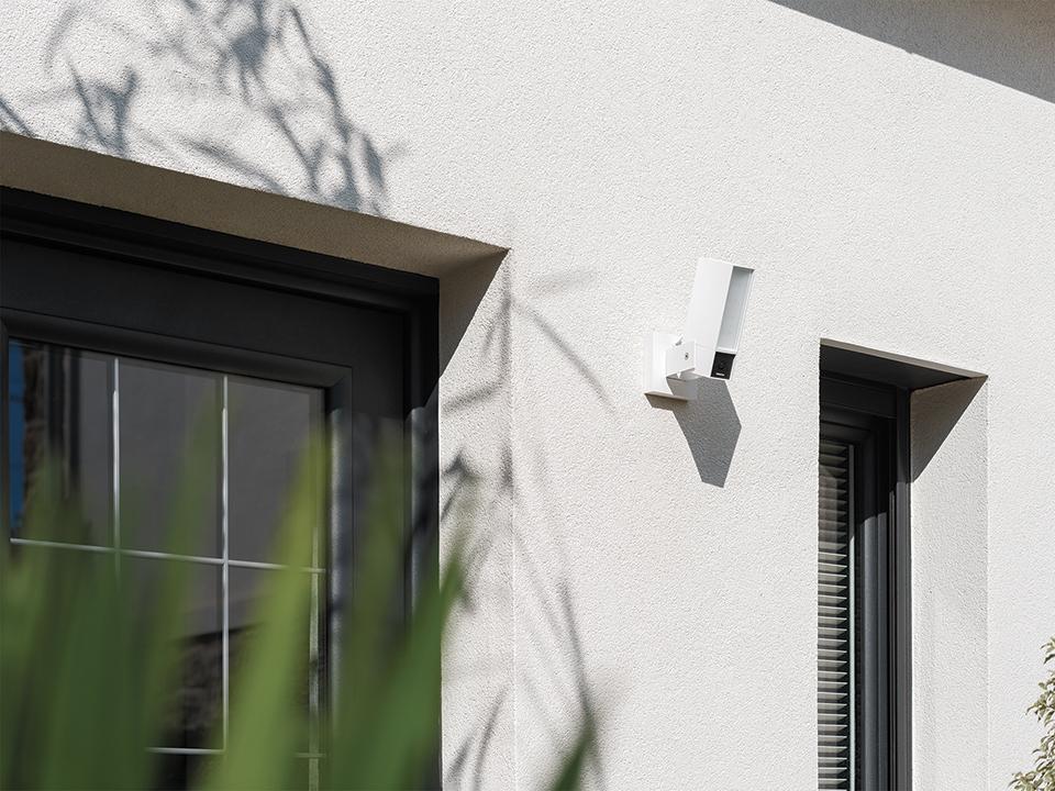 Una telecamera BTicino Outdoor Original bianca è installata all'esterno, tra una porta e una finestra di un edificio moderno.